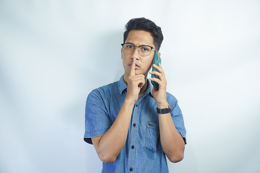 Asian man wearing blue shirt hides a secret while talking on the phone with finger on lips against white background.