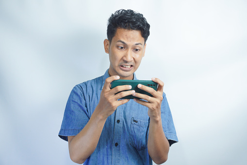 Young Asian man playing a video game on mobile phone and got negative reactions. Facial expressions could be furious, fierce, upset, unhappy, irritated, frustrated.