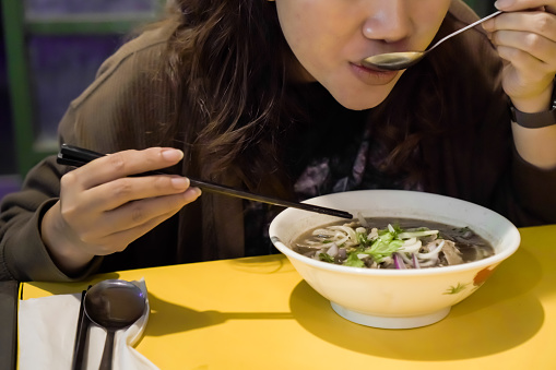 Asian woman eating Vietnamese soup dish Pho that consisting of broth, rice noodles, herbs, and beef pork chicken meat with scallion, onion, cilantro (coriander leaves), bean sprouts, lime, chili oil.