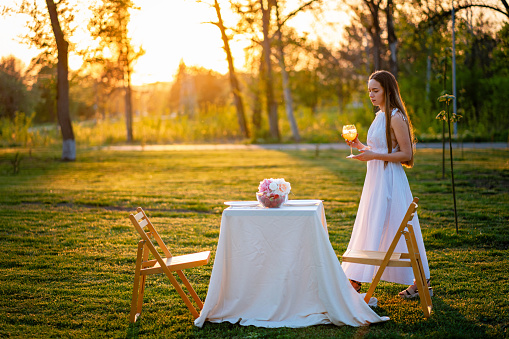 Beautiful woman in a white dress arranging a table for two for a romantic date outdoors during a sunset