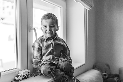Black and white portrait of cheerful little boy sitting on window sill at home