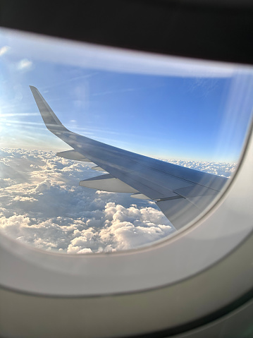 Stock photo showing the wing of a plane pictured through the airplane cabin window with views of blue sky and cumulous clouds.