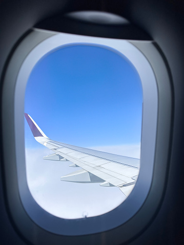 Stock photo showing the wing of a plane pictured through the airplane cabin window with views of blue sky and cumulous clouds.