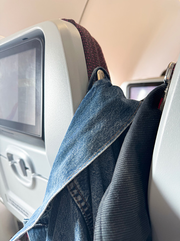 Stock photo showing close-up view of the interior of an airplane with row of two seats in an economy class cabin of a passenger plane. A seat is viewed from the rear with a built-in fold down table and headrest hooks from which denim jackets hang by material loops.