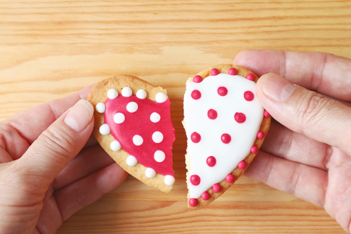 Woman's hand and man's hand holding two half heart shaped cookies attach on wooden background