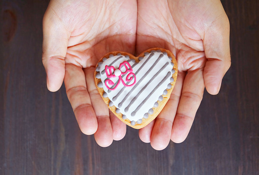Hands holding a heart shaped white chocolate glazed cookie on black wooden background