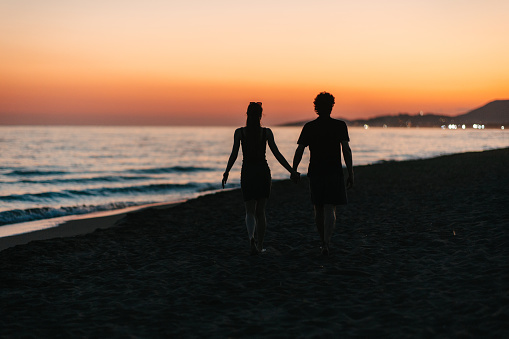 The silhouettes of a happy couple on a romantic walk by the sea at sunset, just before nightfall, holding hands
