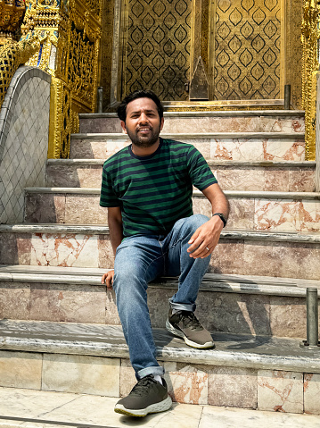 Stock photo showing close-up view of an Indian tourist sitting outside the ornately, decorated walls of Wat Phra Kaeo (Royal Grand Palace), Bangkok, Thailand.