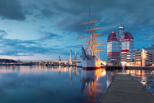Gothenburg, Sweden- April 8, 2023: A historic sailing ship moored at  waterfront, with the iconic Lilla Bommen building illuminated in the evening, concept of maritime heritage and travel.