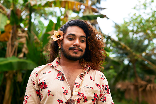 Gay, showing pride, diversity, radiates happiness. Confident man with flower in hair poses in nature. Outdoors, represents inclusion, nature beauty. Floral shirt complements tropical setting.
