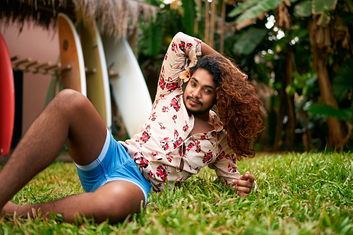 Blue shorts, floral shirt enhance vivid look. Confident gay man with rich curly hair reclines in tropical garden, vibrant style. LGBT travel, self-love ambiance resonate. Leisure in natural, sunny.