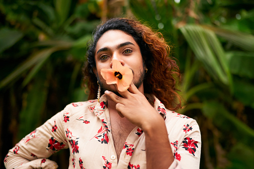 Gender fluid individual beauty, fashion diversity. South Asian man in floral shirt poses with hibiscus flower in mouth, expresses femininity amid green plants. Unique, vibrant portrait outdoors.