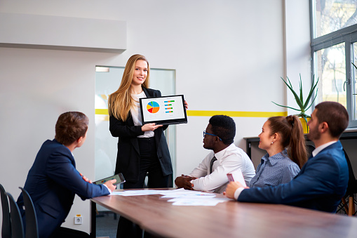 Confident female CEO presents colorful chart on convertible laptop to diverse, multiethnic business team. Pro young leader conducts strategy session, african, asian, caucasian colleagues on a meeting.