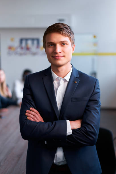Colleagues in background work at meeting. Confident young businessman stands in office, arms crossed, smiling. Professional male wears blue suit, white shirt, no tie, exemplifies leadership. un joven hombre de negocios seguro de sí mismo está de pie en el cargo, con los brazos cruzados, sonriendo. los colegas en segundo plano trabajan en la reunión. el hombre profesional viste traje azul, camisa blanca, sin corbata, ejemplifica el liderazgo