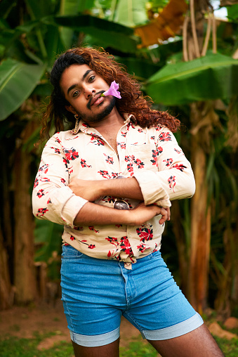 Expresses queer pride, gender fluidity. Confident gay man with curly hair, flower in mouth, stands in tropical garden. Shorts, floral shirt enhance his bold, colorful style amidst green foliage.