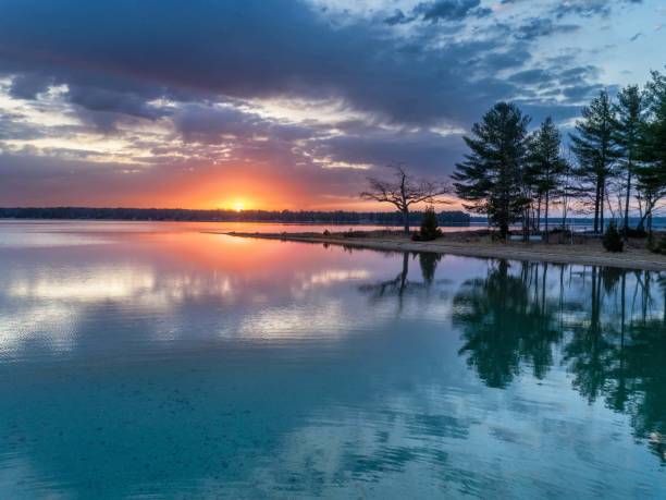 Higgins Lake, Michigan. 4/9/24. A tranquil lake reflects the vibrant hues of sunset, with broad streaks of pink and orange light breaking through the cloud cover. A tree-lined shoreline gently meanders into the distance on the right, providing a stark contrast to the open waters. Drone point of view.