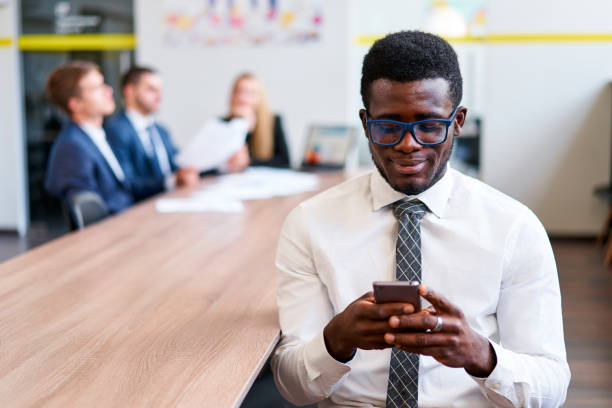 istock African businessman in glasses uses smartphone in modern office. Focused on screen, tech tool at work. Team discusses project in background. Mobile corporate communication, efficiency in foreground. 2148602348