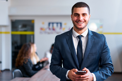 Colleagues work in background. Smartly dressed eastern businessman smiles, holding smartphone in modern office. Pro uses fintech app for efficient business management and transactions.