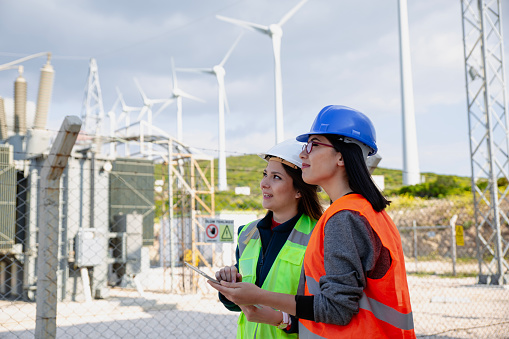Two young electrical female engineers with protective workwear standing, working, checking conducts with digital tablet pc in front of electrical components, wind turbines and towers of high tension of an thermal electricity power generation station field