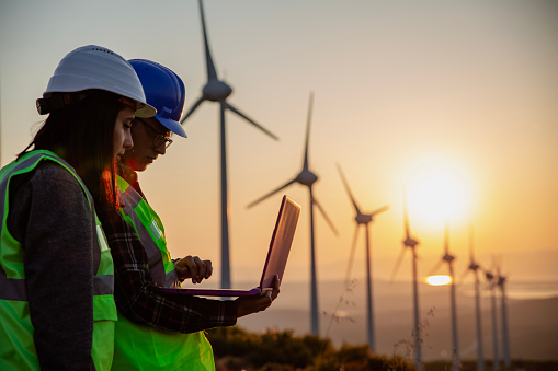Young electrical engineer woman and business man standing in front of wind turbines checking and working about technical problems and writes the results of measurements with laptop pc in wind power plant electric energy station.