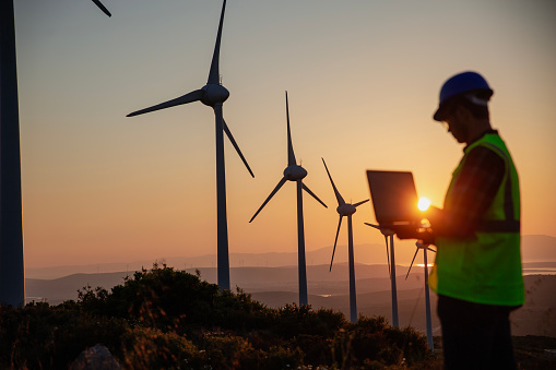 Silhouette of a young male engineer holding a laptop computer planning and working for the energy industry and standing beside a wind turbines farm power station at sunset time.