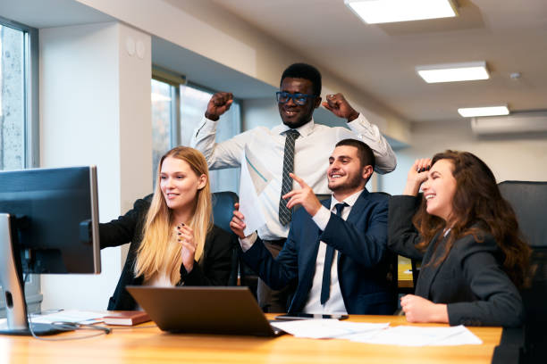 istock Multiethnic office team rejoices around computer, happy African exec raises fists in victory, coworkers cheer, man points at screen showing growth chart, workplace celebration of successful deal. 2148602994