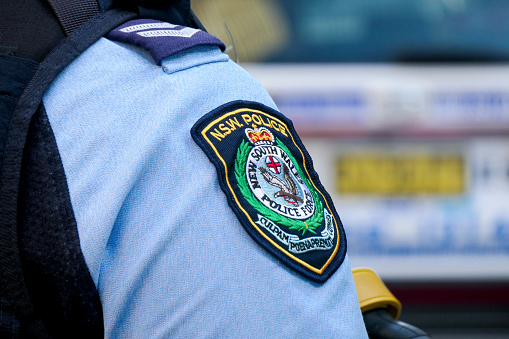 The shoulder emblem and rank stripes of an officer of the New South Wales Police Force standing on the street. Their taser gun with the yellow holster is visible on the right. In the background is a police vehicle.  This image was taken at the corner of Oxford Street and Hollywood Avenue, Bondi Junction on a sunny afternoon on 14 April 2024.