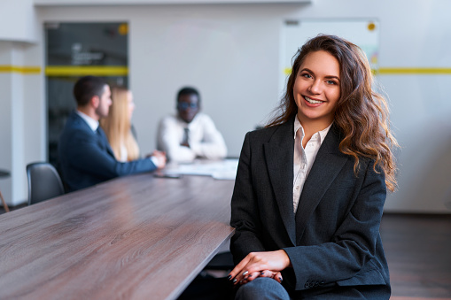 Colleagues discuss in background at office meeting. Confident young businesswoman with curly hair, wearing a suit, smiles seated at boardroom table. Leadership, team, ambition, success themes.