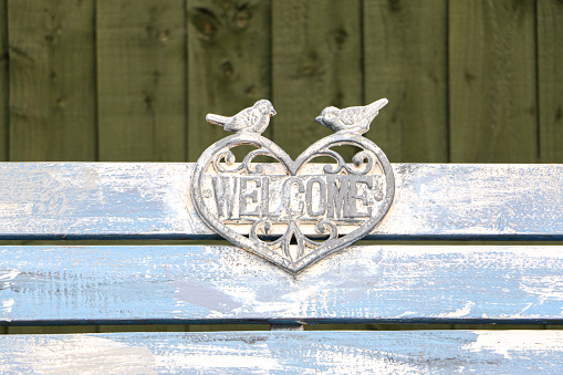 Welcome sign designed in heart shape with two birds on top, on a park bench which is light blue but losing colour