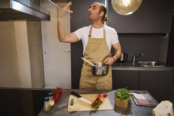 Handsome male chef holding saucepan with tomato sauce, kissing fingers, showing tasty hands sign, saying bellissimo cooking gourmet food in the home kitchen. People. Culinary. Alimentation. Lifestyle. chef masculino guapo sosteniendo una cacerola con salsa de tomate, besando los dedos, mostrando un sabroso signo de manos, diciendo bellissimo cocinando comida gourmet en la cocina de la casa. gente. culinario. alimentación. modo de vida