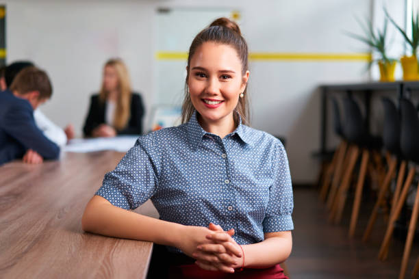 Positive, pro ambiance, teamwork in contemporary workplace, career woman focus. Confident Asian woman in smart casual attire sits at office meeting table, colleagues in discussion behind her. una mujer asiática segura de sí misma con un atuendo informal elegante se sienta en la mesa de reuniones de la oficina, con colegas en discusión detrás de ella. ambiente positivo y profesional, trabajo en equipo en el lugar de trabajo contemporáneo, e