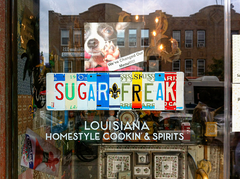 On a glass entry door, a sign reads Sugar Freak in what looks like a multi-colored drivers license plate. Under the plate, the lettering reads Louisiana Homestyle Cookin' & Spirits. Through the glass, eclectic decor can be seen. And in the door reflection, other New York City buildings and people walking by are reflected framing the signage.