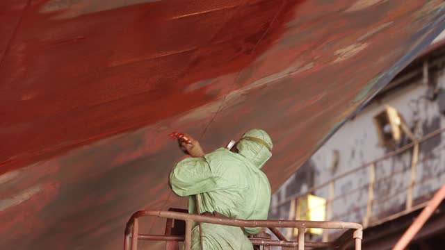 Worker in protective gear applies red paint to ship hull in dry dock. Industrial painting, maintenance against rust, maritime vessel refurbishment process. Shipyard worker, safety equipment in use.