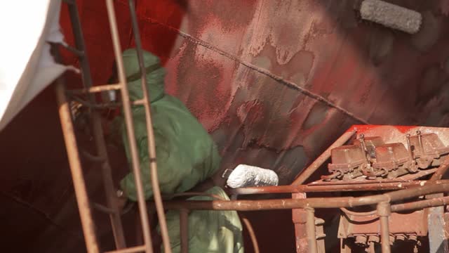 Worker in protective gear paints hull in shipyard. Industrial maritime maintenance, ship renovation process. Manual labor, protective equipment, vessel restoration by craftsman.