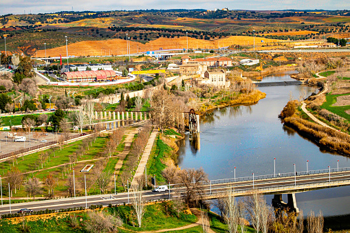 Aerial view of valley with Tagus river on outskirts of Toledo city, hills with agricultural land in background, vehicle bridge over river, buildings, water mill, industries, sunny day in Spain