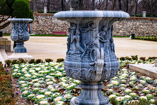 Closeup of French vases in Jardín del Parterre in Retiro park, decorated with a goat head, esplanade, stone wall and bare tree trunks in background, cloudy day in Madrid, Spain
