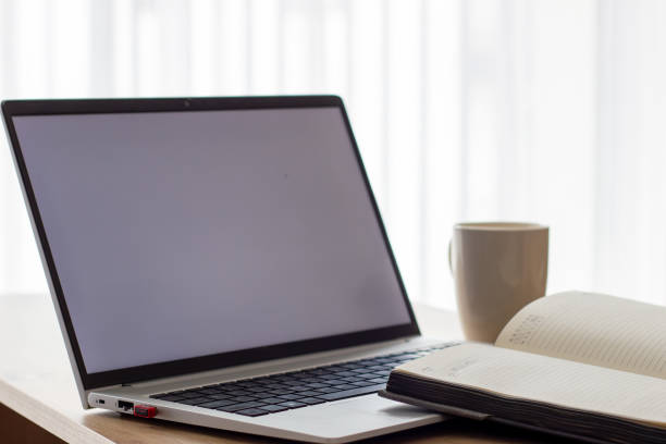 notebook in front of a laptop with blank white screen, at a small table, image in light colors, blurred background and white cup