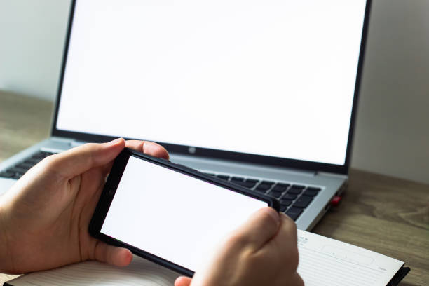 blank white screens of mobile phone and laptop, man holding phone in front of laptop at the table, scrolling , notebook and pen nearby, procrastination at work