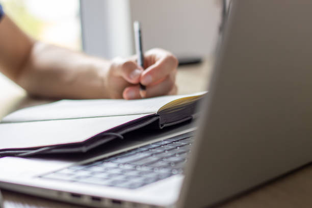 istock laptop in focus, man on background makes notes in paper notebook, home office 2148608837
