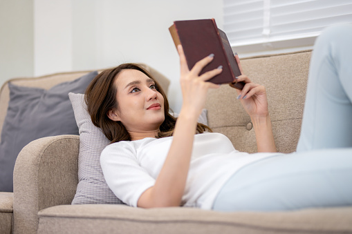 A woman enjoys reading a hardcover book, lying comfortably on a couch in a well-lit living room.
