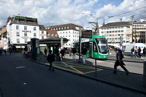 Basel, Switzerland - March 21, 2024: Basel City Public Transportation Tram Arrival Departure Platform, Building Exterior, Retail Store, Land Vehicle, People Walking, Waiting For Tram, Departing After Arrival With Tram Scene And More During Springtime