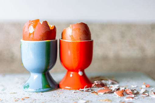 The aftermath of a plate of boiled eggs for breakfast. Two empty and cracked eggshells sit in their respective blue and orange egg cups, with fragments of eggshell on the surrounding chopping board.