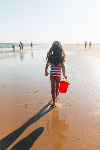 Cheerful little girl running and playing with red bucket, splashing with water from the ocean on the beautiful beach area in Algarve Portugal