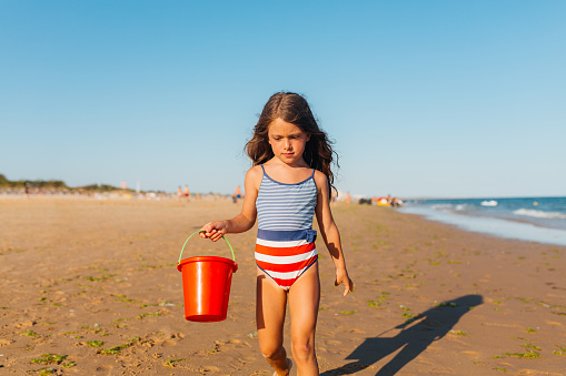 Cheerful little girl running and playing with red bucket, splashing with water from the ocean on the beautiful beach area in Algarve Portugal