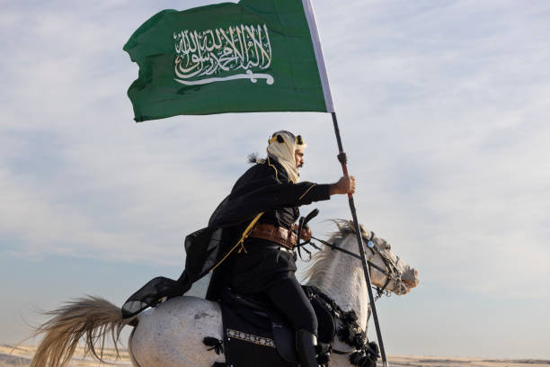 Saudi man in traditional clothing in the deset with a white horse Man in traditional clothing with his horse in a desert, riding with Saudi flag in his hand arab man riding horse stock pictures, royalty-free photos & images