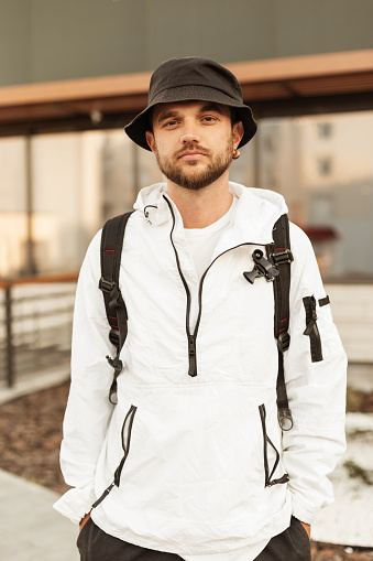 Stylish young man hipster with trendy hat with white urban jacket and backpack in the city