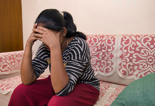 A woman sits alone on a couch, her hands covering her face in a gesture of despair.