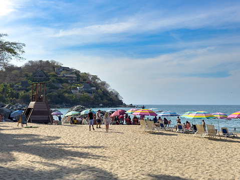 SAYULITA, NAYARIT, MEXICO - 2024-04-13: Vacationers and local beachgoers enjoy the sun, sea, and sand at public beach: Playa Los Muertos in Sayulita, with a vibrant display of colourful umbrellas lining the shore, nestled against a backdrop of lush hills with quaint houses, capturing the quintessential relaxed atmosphere of a sunny afternoon on the Pacific coast.