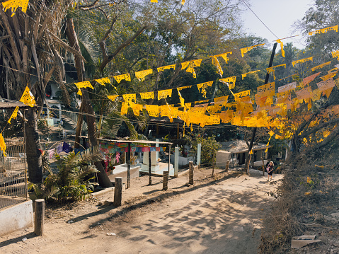 SAYULITA, NAYARIT, MEXICO - 2024-04-10: Along the cemetery road adjacent to Playa Los Muertos in Sayulita, Nayarit, Mexico, vibrant yellow papel picado banners flutter in the breeze, creating a festive atmosphere that guides visitors and locals alike through a path rich with cultural symbolism, set against a backdrop of tropical vegetation and clear blue skies.