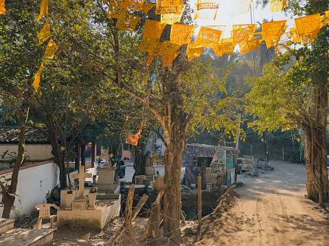 SAYULITA, NAYARIT, MEXICO - 2024-04-10: The warm, golden hour sunlight filters through the trees along the cemetery road next to Playa Los Muertos in Sayulita, Nayarit, Mexico, casting a serene glow on the papel picado that adorns the route, with gravestones resting peacefully amidst the living landscape, a testament to the town’s rich cultural tapestry.